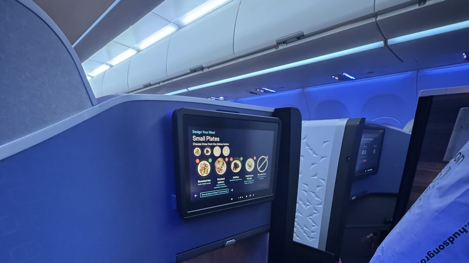 JetBlue Mint Suite cabin on the A321LR from JFK to London — seatback screen showing the meal-design menu, blue mood lighting, and the private suite shell