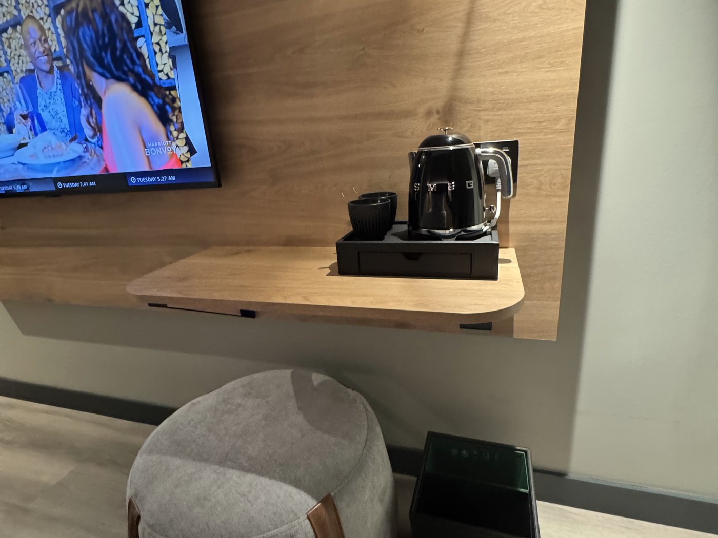 Moxy Piccadilly Circus room details — floating wood desk with a black SMEG kettle, coffee cups, wall-mounted TV, and a felt floor pouf