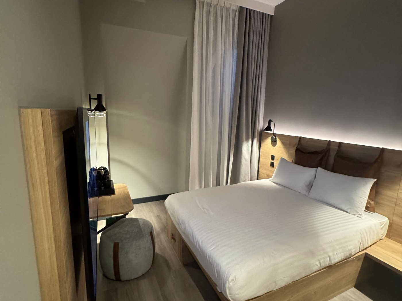 Inside a compact Moxy London Piccadilly Circus room — queen bed with wood headboard, reading lights, wall-mounted TV, and a floor pouf